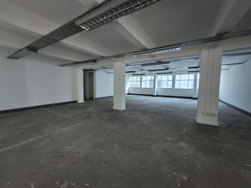 A spacious, open-plan office area featuring industrial-style exposed ceiling beams, fluorescent lighting, and concrete flooring with large windows providing natural light.