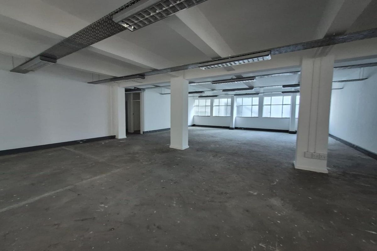 A spacious, open-plan office area featuring industrial-style exposed ceiling beams, fluorescent lighting, and concrete flooring with large windows providing natural light.