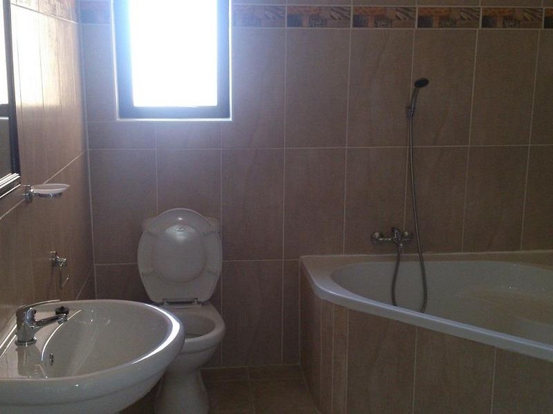 A functional bathroom featuring a corner bathtub with handheld shower, pedestal washbasin, toilet, and neutral wall tiling with a decorative border.