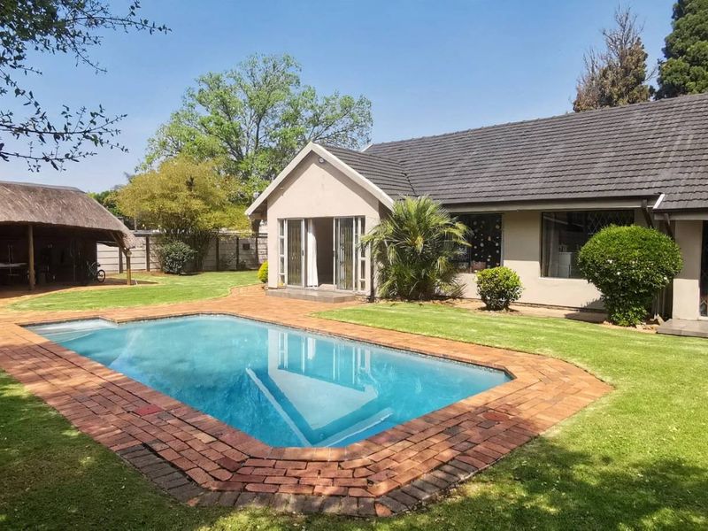 Rear garden view featuring a swimming pool, brick-paved surround, and a detached thatched lapa, set against the rear facade of the house.