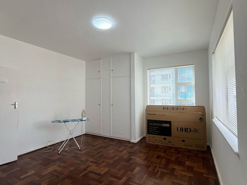 A bright, empty bedroom featuring classic parquet flooring, built-in storage cupboards, and large windows fitted with venetian blinds.