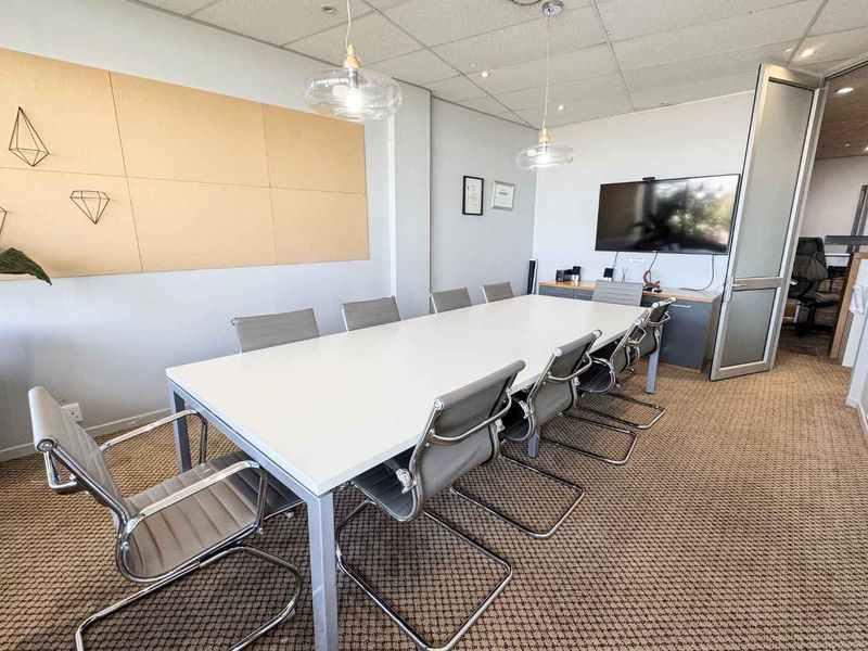 A professional boardroom or meeting room featuring carpeted flooring, suspended ceiling with integrated lighting, and a glass-panelled entrance door.