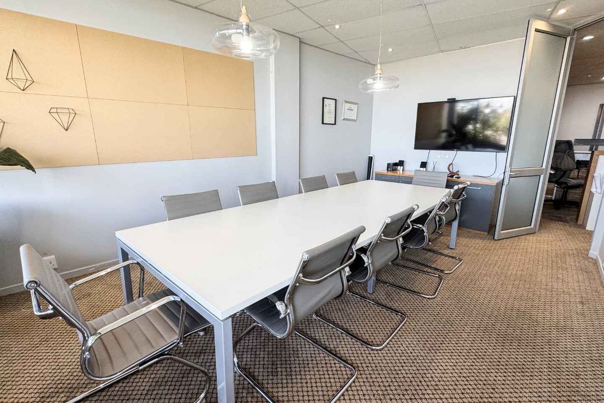 A professional boardroom or meeting room featuring carpeted flooring, suspended ceiling with integrated lighting, and a glass-panelled entrance door.