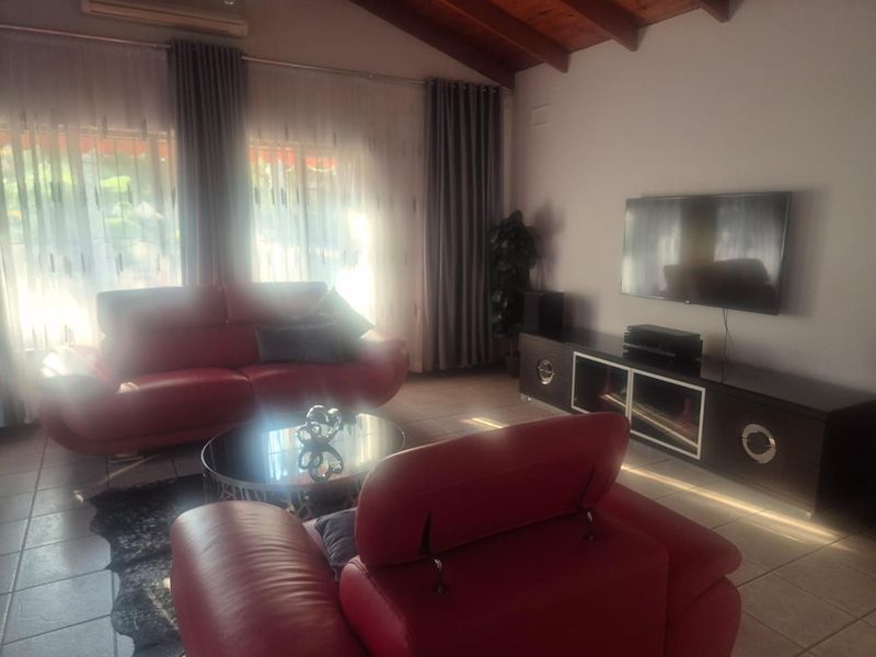 A living room featuring exposed timber ceiling beams, tiled flooring, and a wall-mounted television, with large windows providing natural light.