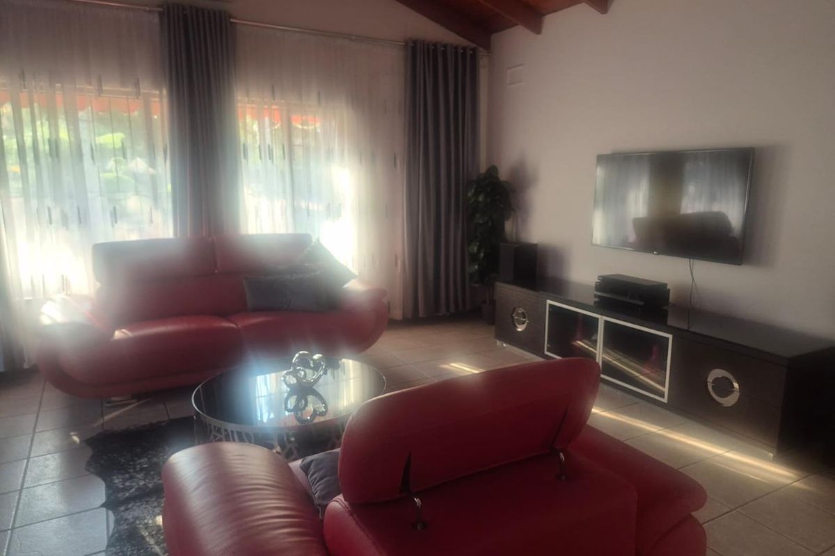A living room featuring exposed timber ceiling beams, tiled flooring, and a wall-mounted television, with large windows providing natural light.