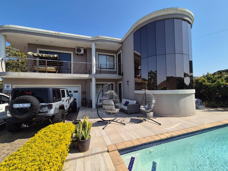 Rear exterior view featuring a swimming pool, tiled patio area, curved glass architectural feature, and a balcony with stainless steel railings.