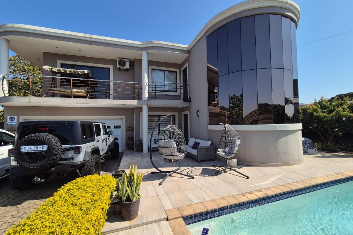 Rear exterior view featuring a swimming pool, tiled patio area, curved glass architectural feature, and a balcony with stainless steel railings.