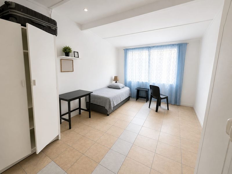 A functional bedroom featuring neutral tiled flooring, recessed ceiling lighting, and a built-in wall shelf, offering a clean and simple living space.
