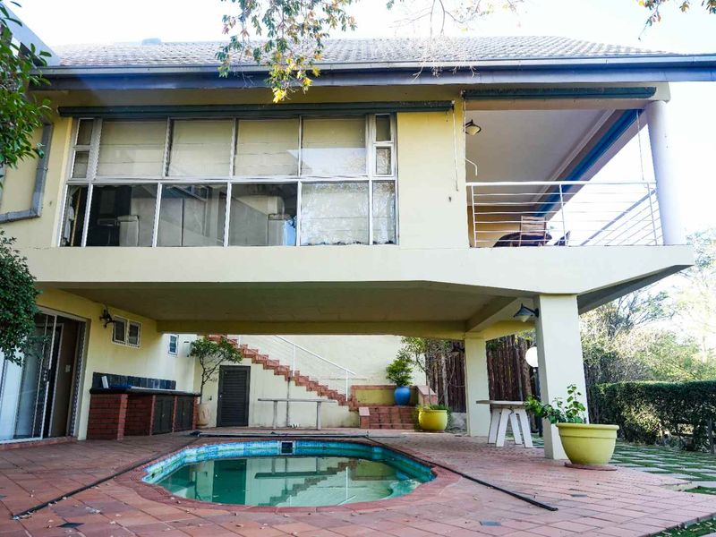 Rear exterior view featuring a swimming pool, paved patio area, built-in braai, and an elevated balcony overlooking the garden.