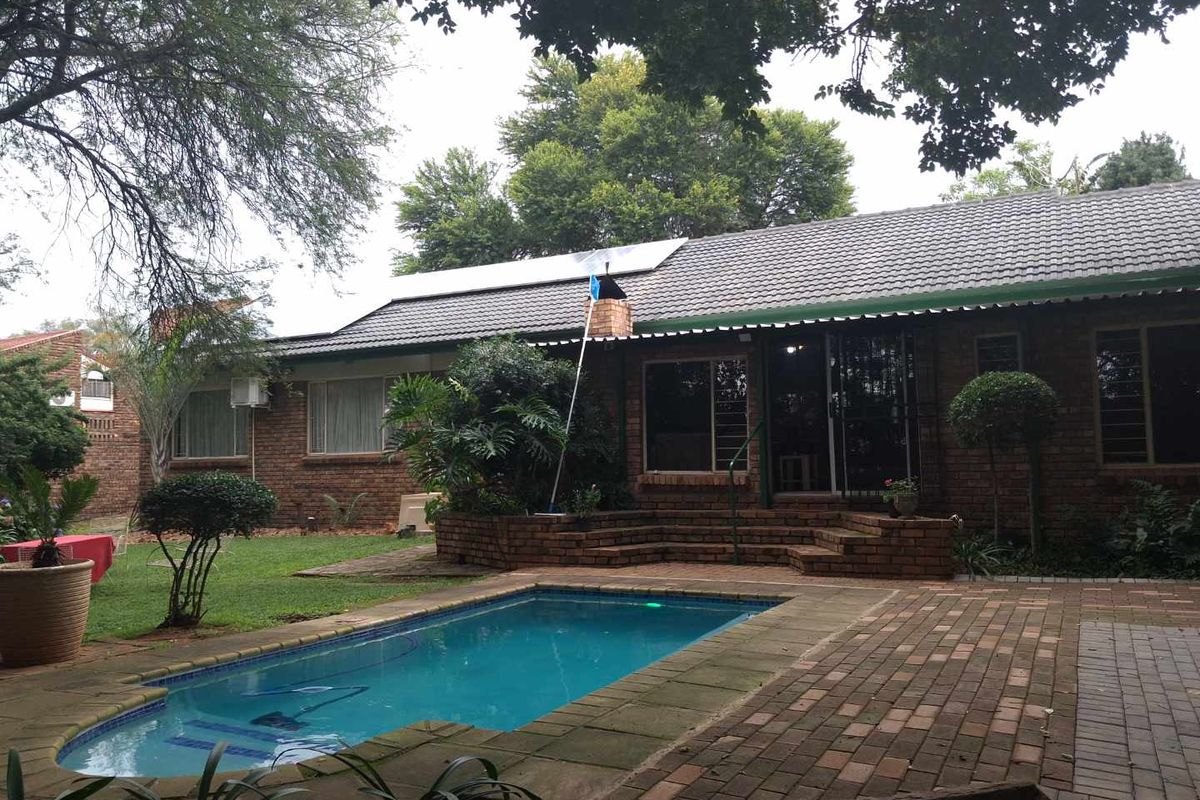 Rear view of a brick house featuring a swimming pool, paved patio area, and a landscaped garden with mature trees.