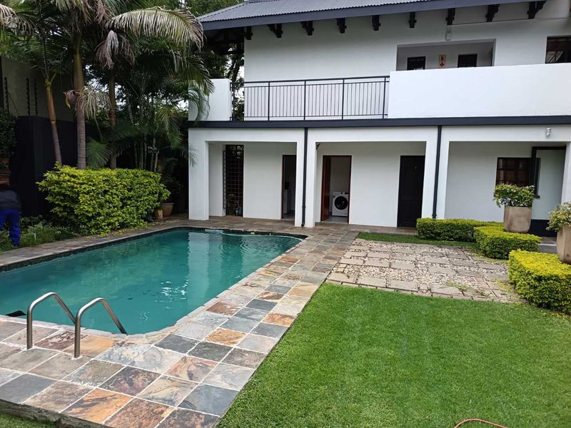 A well-maintained outdoor pool area featuring stone paving, a manicured lawn, and a two-story building with a balcony and metal railing.