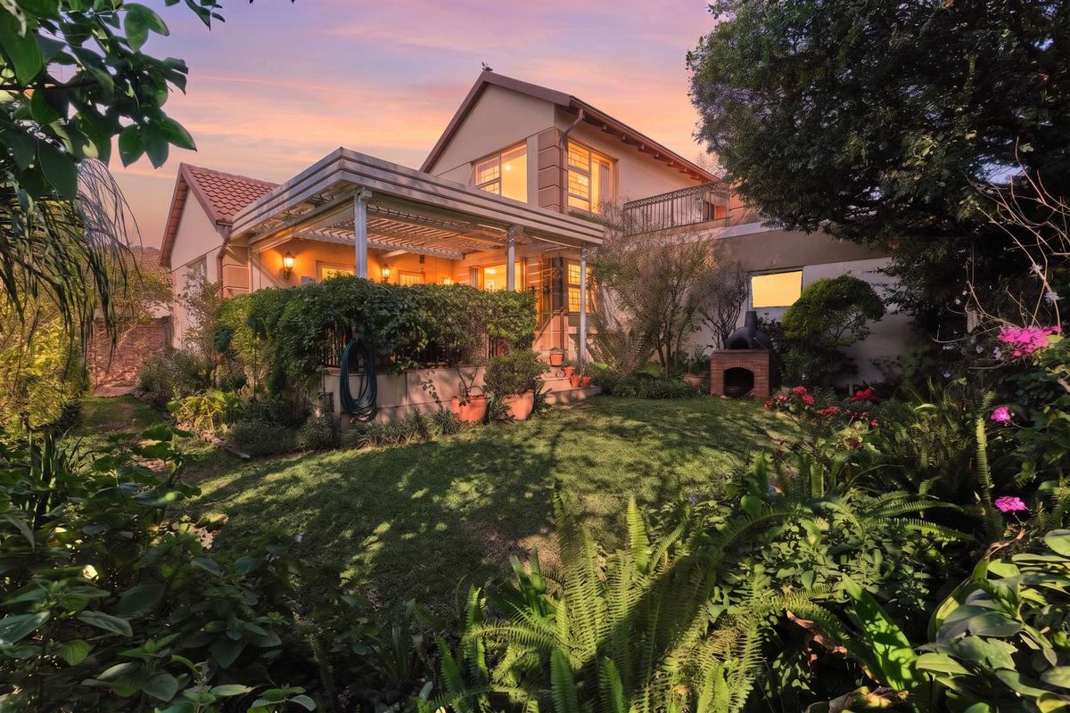 A beautiful rear view of the property featuring a lush landscaped garden, a covered patio area, and a built-in outdoor braai.