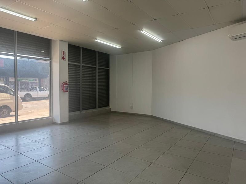 A bright, open-plan retail space featuring large glass storefront windows, tiled flooring, suspended ceilings, and a wall-mounted air conditioning unit.