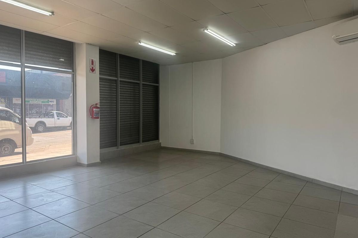 A bright, open-plan retail space featuring large glass storefront windows, tiled flooring, suspended ceilings, and a wall-mounted air conditioning unit.