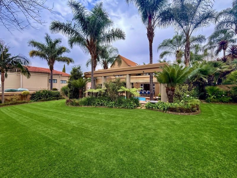 A well-maintained rear garden featuring a lush lawn, mature palm trees, and a covered patio area overlooking a swimming pool.