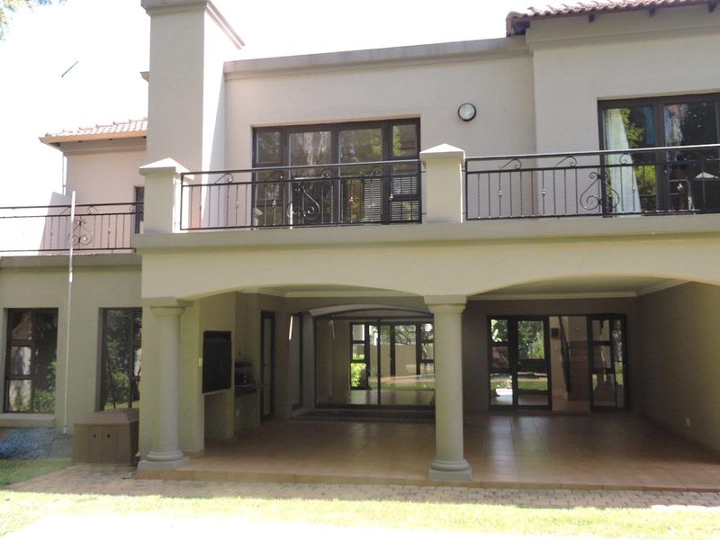 Rear elevation featuring a spacious covered patio with tiled flooring, columns, and a built-in braai, leading to an upper-level balcony with wrought iron railings.