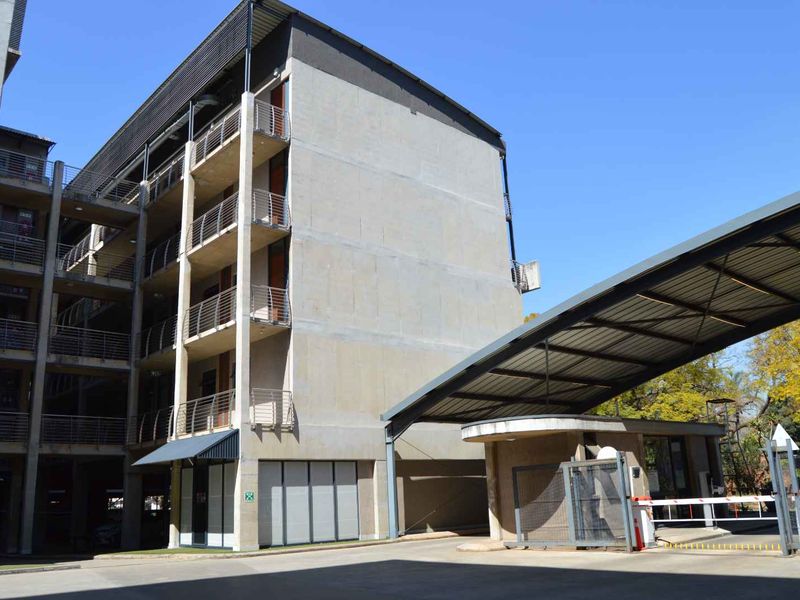 Exterior view of the apartment complex entrance featuring a security boom gate, guard house, and multi-story building with concrete facade and balcony railings.