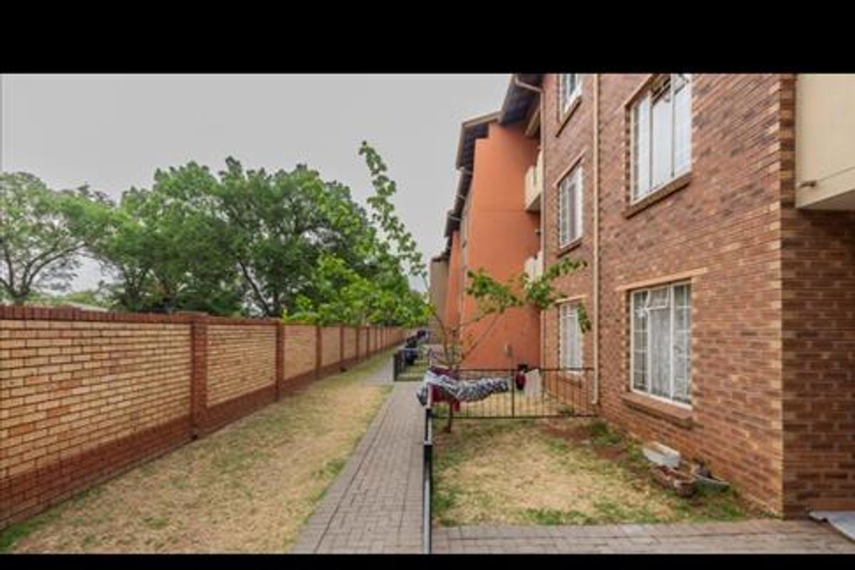 Exterior view of the apartment building showing a paved walkway, brick boundary wall, and communal garden space.