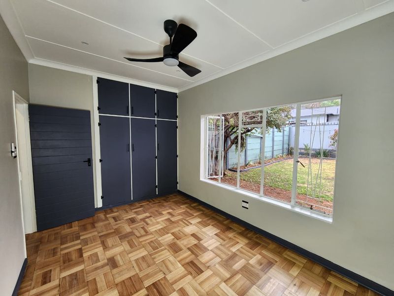 A bright, empty bedroom featuring polished parquet flooring, dark built-in cupboards, a modern ceiling fan, and large windows overlooking the garden.