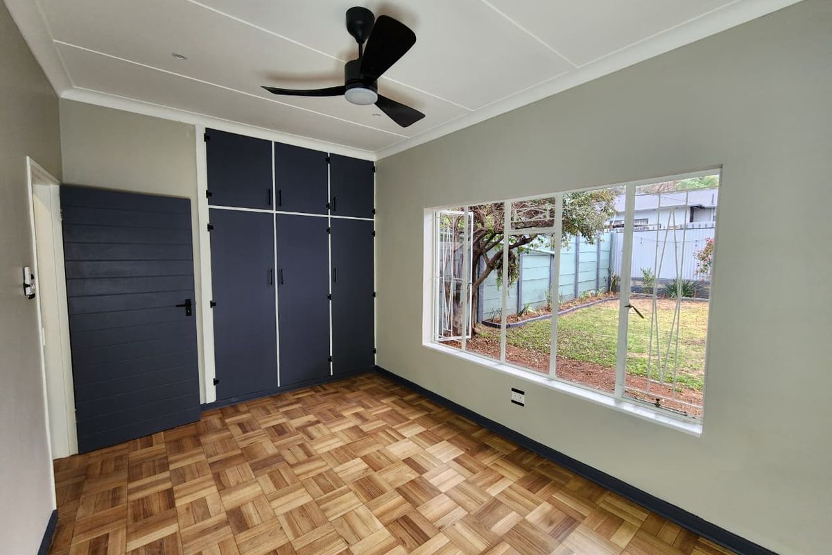 A bright, empty bedroom featuring polished parquet flooring, dark built-in cupboards, a modern ceiling fan, and large windows overlooking the garden.