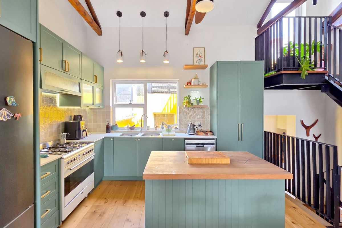 Modern kitchen featuring sage green cabinetry, timber island countertop, exposed ceiling beams, and high-quality finishes, overlooking an open-plan living area with mezzanine access.
