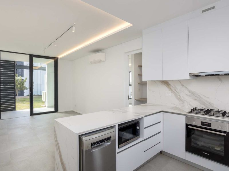 Modern open-plan kitchen and living area featuring sleek white cabinetry, stone countertops, integrated appliances, and direct access to a private outdoor patio.
