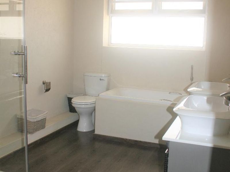 A modern bathroom featuring a glass shower enclosure, built-in bathtub, dual vanity basins, and dark laminate flooring with ample natural light.