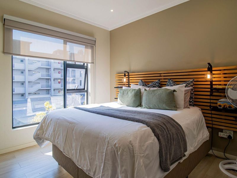 A well-lit bedroom featuring a custom timber slat headboard, laminate flooring, and a large window with a roller blind.