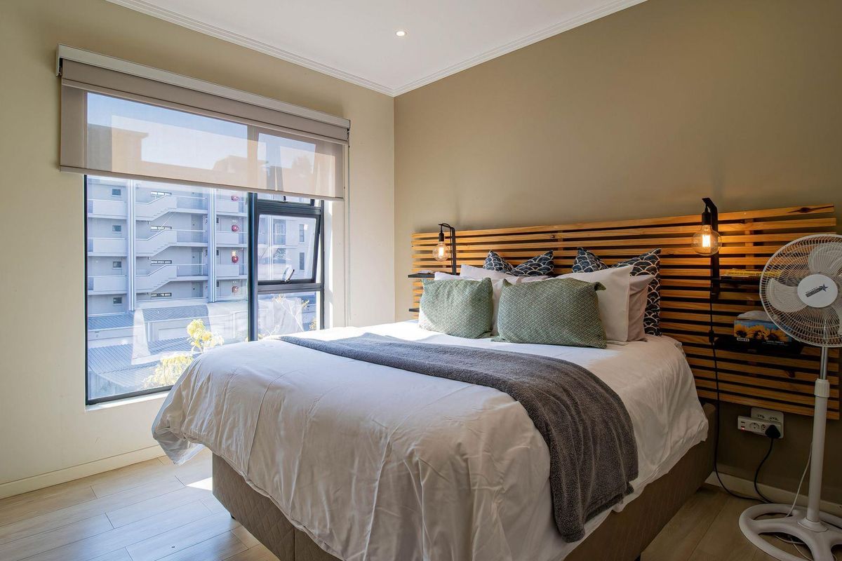 A well-lit bedroom featuring a custom timber slat headboard, laminate flooring, and a large window with a roller blind.