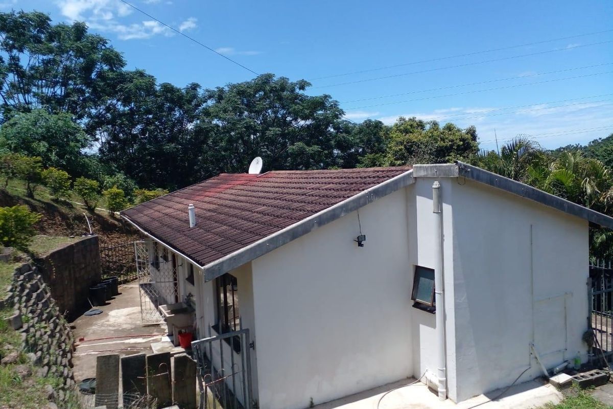 Rear view of the property showing the tiled roof, exterior walls, concrete apron, and surrounding garden area with a retaining wall.