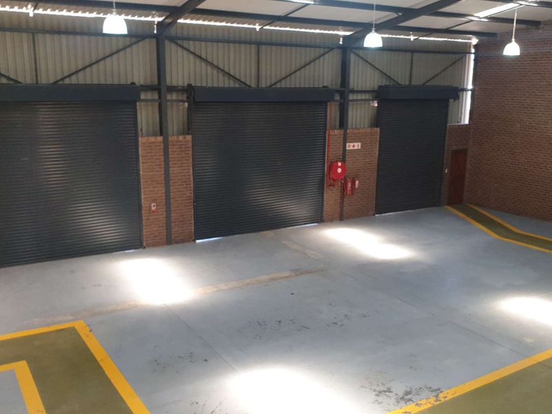 Spacious industrial warehouse or garage featuring high ceilings with exposed steel trusses, multiple roller shutter doors, and durable painted concrete flooring.