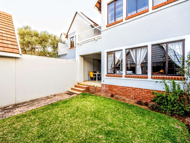 Rear view of the townhouse featuring a private garden, paved walkway, covered patio area, and upper-level balcony with brick and plaster finishes.