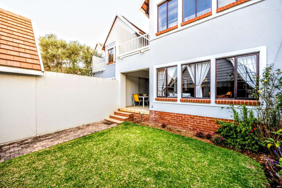 Rear view of the townhouse featuring a private garden, paved walkway, covered patio area, and upper-level balcony with brick and plaster finishes.