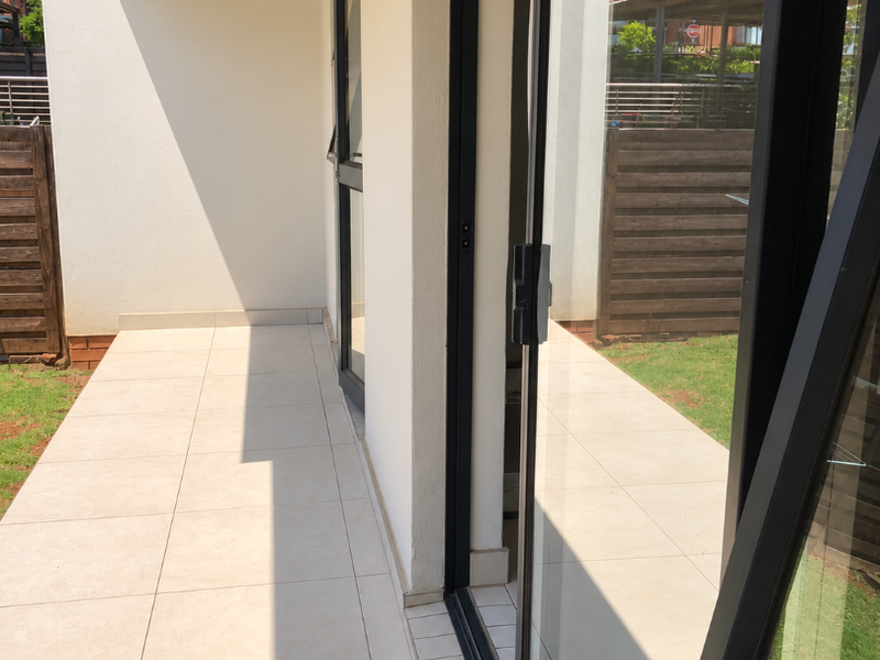 A clean, modern covered patio featuring light-toned floor tiles, black-framed sliding glass doors, and a private timber screen overlooking a small garden area.