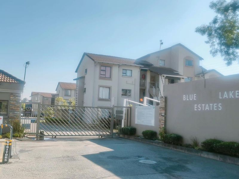 Front entrance view of Blue Lake Estates apartment complex featuring a secure gated access point, perimeter wall, and multi-story residential buildings.