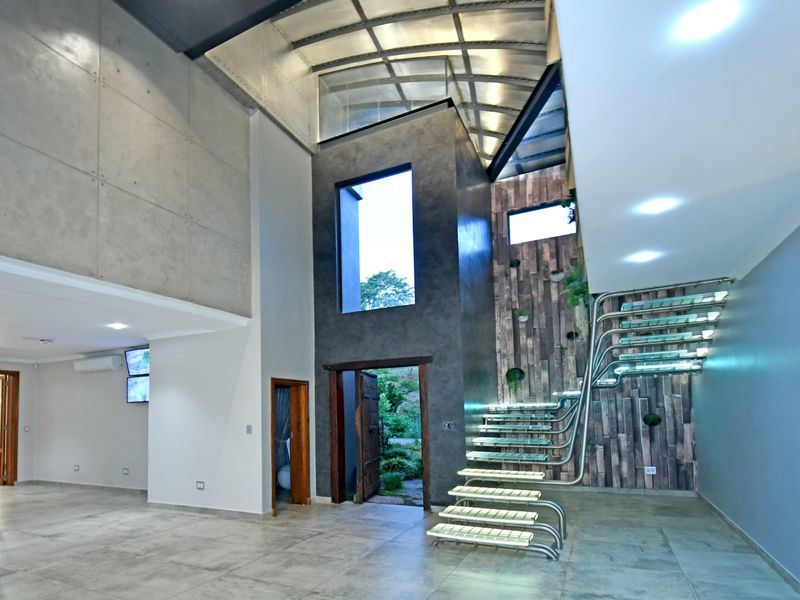 Spacious modern living area featuring a striking floating glass staircase, high vaulted ceilings, exposed concrete wall panels, and premium tiled flooring.