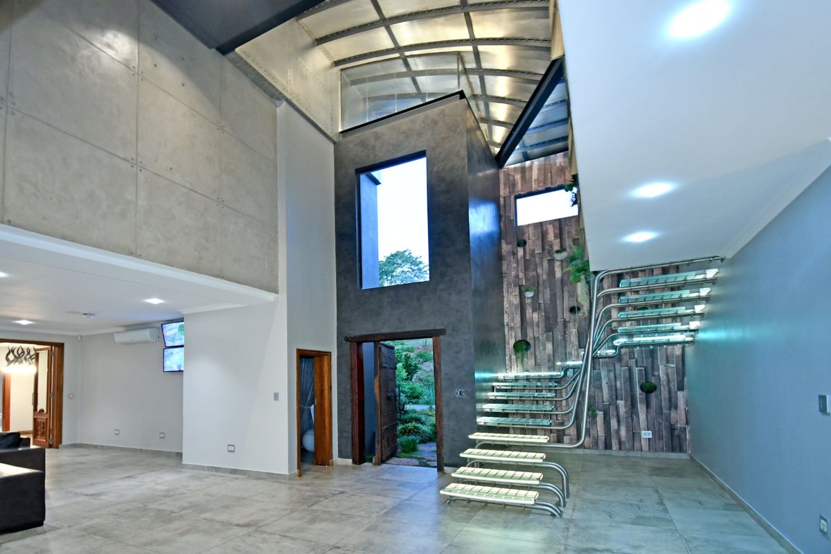 Spacious modern living area featuring a striking floating glass staircase, high vaulted ceilings, exposed concrete wall panels, and premium tiled flooring.