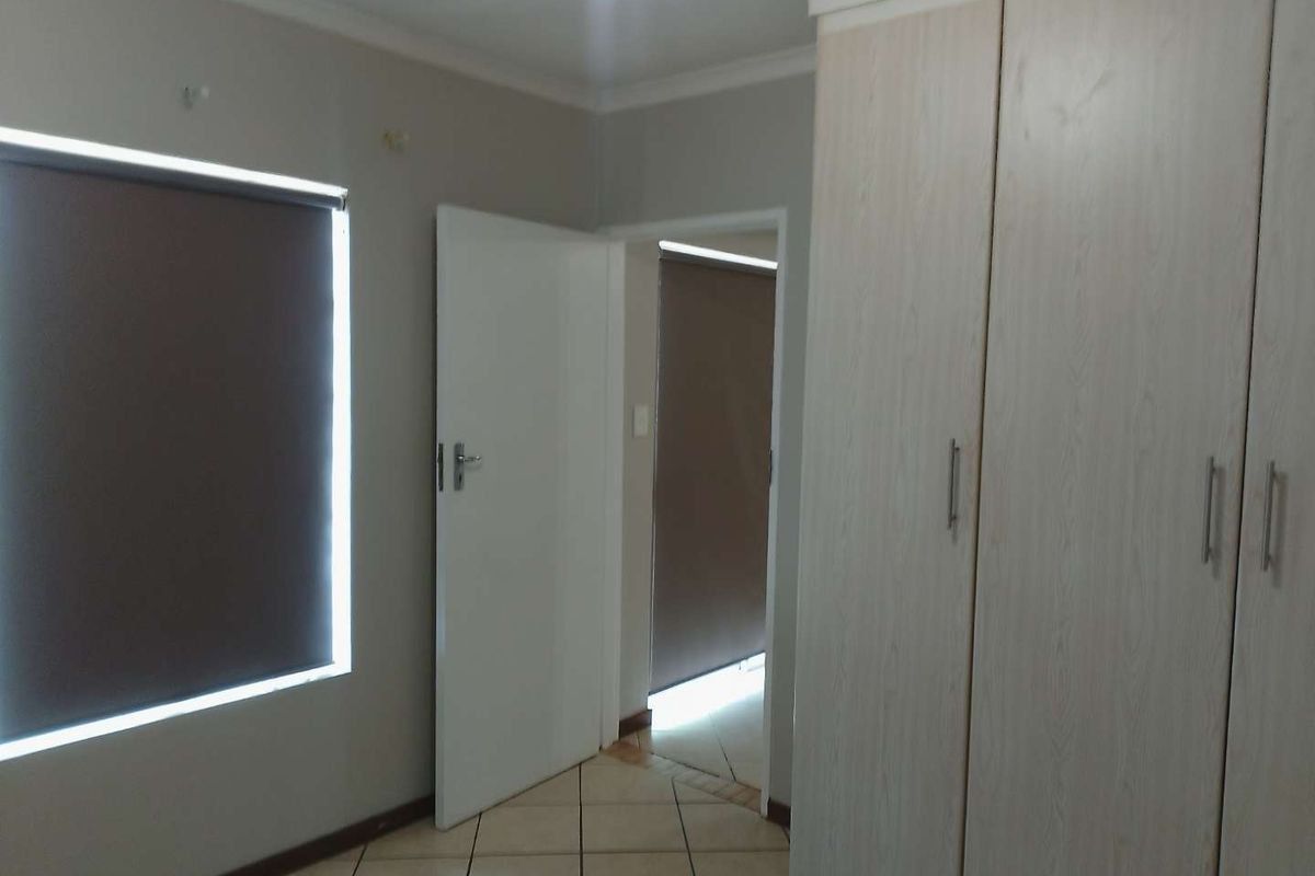 A clean, empty bedroom featuring neutral walls, tiled flooring, and built-in cupboards with roller blinds on the windows.