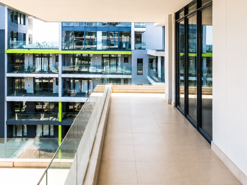 A spacious, modern covered balcony featuring large-format floor tiles, a sleek glass balustrade, and expansive sliding glass doors leading into the apartment.