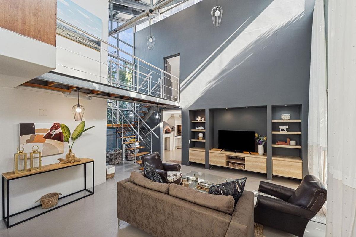 Spacious double-volume living area featuring a mezzanine level, industrial-style staircase, built-in shelving, and large windows providing abundant natural light.