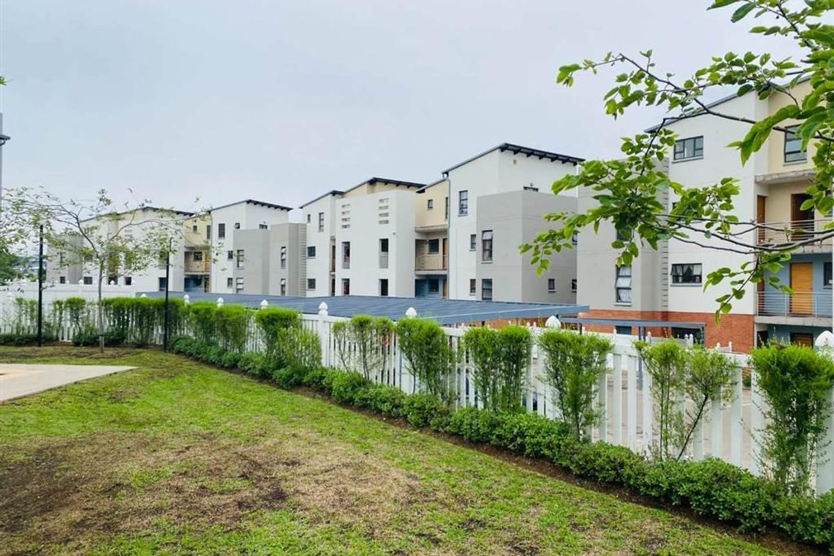 View of the rear exterior of a modern apartment complex featuring a white picket fence, landscaped hedges, and paved walkways.