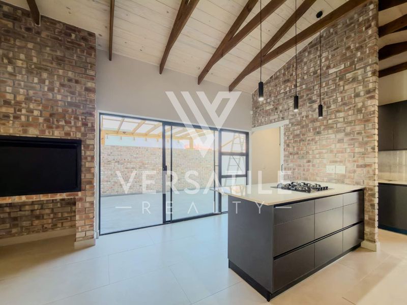 Modern open-plan living area featuring exposed brick walls, vaulted ceilings with timber beams, a central kitchen island with gas hob, and sliding door access.