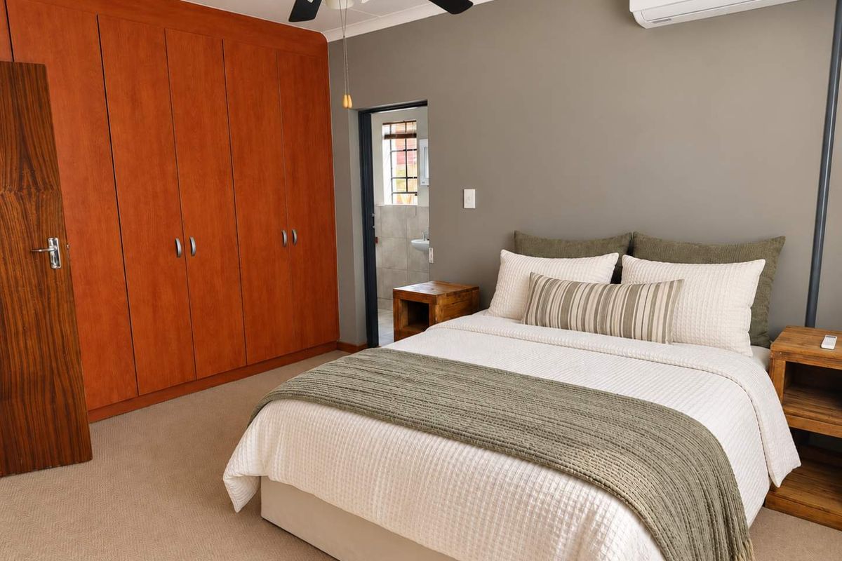 A well-maintained bedroom featuring extensive built-in wooden cupboards, neutral carpeting, a ceiling fan, and a wall-mounted air conditioning unit.