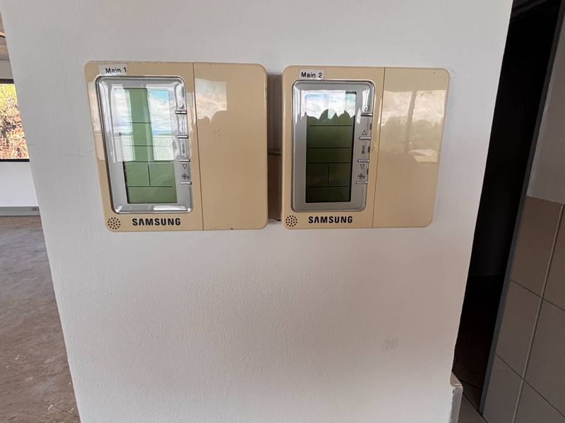 A view of dual Samsung HVAC control panels mounted on a wall within an open-plan office space featuring a suspended ceiling.