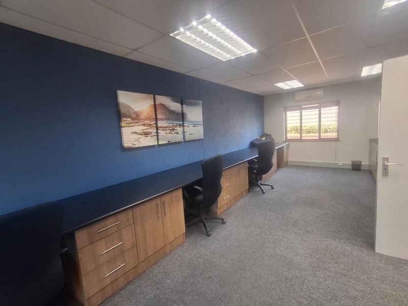 A functional office space featuring a long built-in desk with integrated cabinetry, carpeted flooring, suspended ceiling, and a wall-mounted air conditioning unit.
