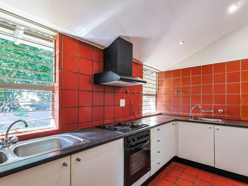 A functional kitchen featuring built-in cabinetry, a stainless steel double sink, electric hob, oven, extractor fan, and distinctive red tiled splashbacks and flooring.