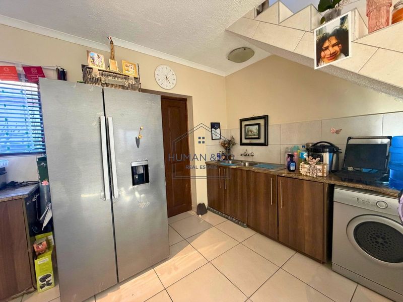 A functional kitchen area featuring built-in cabinetry, a double sink, tiled backsplash, and tiled flooring, situated beneath a staircase.