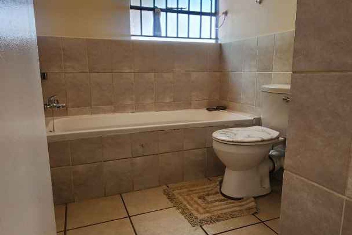 A functional bathroom featuring a built-in bathtub, toilet, and tiled finishes with a security-barred window.