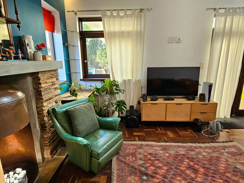 A cozy living room featuring a distinctive stone-clad fireplace, warm parquet flooring, and large timber-framed windows that provide natural light and garden views.
