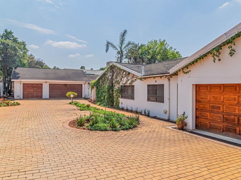Spacious front exterior featuring a large brick-paved driveway, triple garage with wooden doors, and established garden beds.